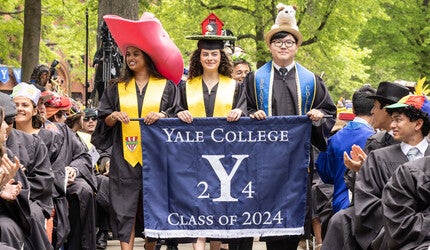 Students wearing festive hats processing with the Yale College Class of 2024 banner.