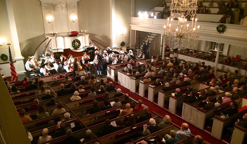 Photo of Orchestra New England, in 18th-century finery, performing a concert.