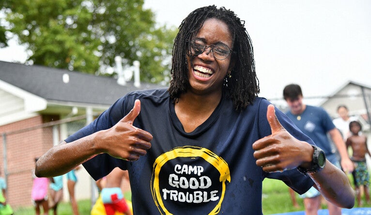 A woman with thumbs up and a t-shirt that says "Camp Good Trouble"