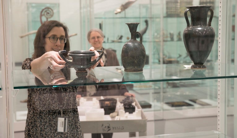 A curator takes an object off a glass shelf while another looks on.