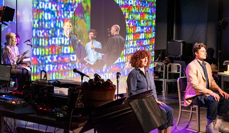 Two performers sit on chairs in front of a scrim with colorful projections. A choir stands behind it, singing.